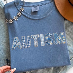 May include: A blue t-shirt with the word "AUTISM" in patchwork letters. The letters feature floral, gingham, and daisy patterns. A silver necklace with flower accents and a brown hat rest on blue jeans.