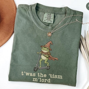 May include: Moss green t-shirt with a graphic of a frog wizard riding a scooter, wearing a hat and cape. The text "t'was the 'tism m'lord" is printed below the graphic. A gold necklace is draped across the shirt.