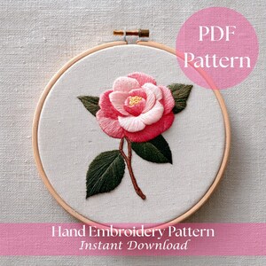 Könnte beinhalten: Ein handgesticktes Blumenmuster in einem Holzreifen. Das Design zeigt eine rosa und weiße Kamelienblüte mit grünen Blättern und einem braunen Stiel. Der Text "PDF Pattern" und "Hand Embroidery Pattern Instant Download" sind sichtbar.