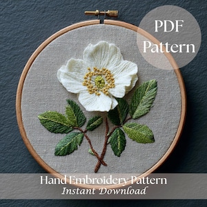 May include: A hand-embroidered floral pattern in a wooden hoop. The design features a white flower with yellow and green details, green leaves, and brown stems on a gray fabric. Text reads "PDF Pattern" and "Hand Embroidery Pattern Instant Download."