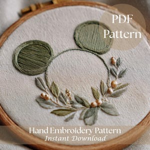 Puede incluir: Patrón de bordado a mano en un aro de madera con un diseño floral verde y una forma de Mickey Mouse. El diseño incluye hojas, bayas y el texto "PDF Pattern" y "Hand Embroidery Pattern Instant Download."