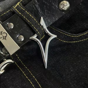 Può includere: Primo piano di un ciondolo argentato a forma di stella appeso a una cintura di pelle nera. La cintura è attaccata a jeans di denim scuro con cuciture gialle. Sono visibili anche una fibbia e rivetti argentati.