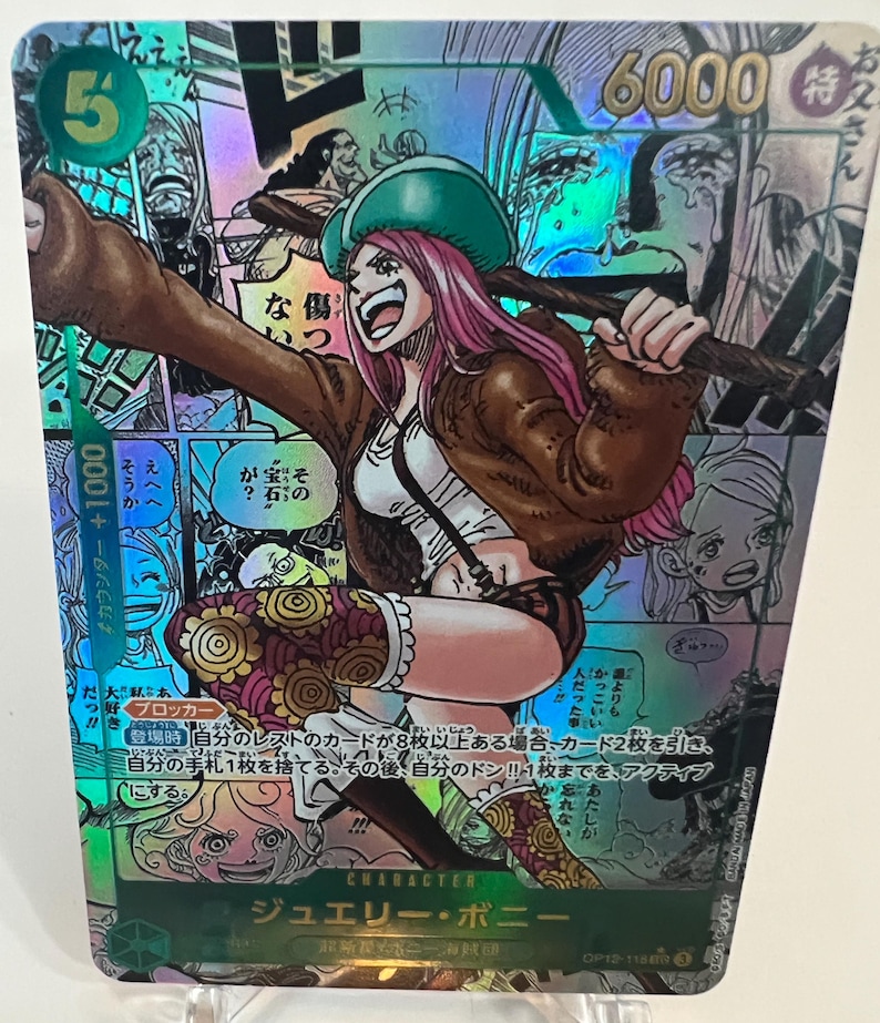 Puede incluir: Carta coleccionable hologr&aacute;fica con Jewelry Bonney de One Piece. La carta tiene un fondo verde con texto japon&eacute;s y una ilustraci&oacute;n de un personaje de una mujer con cabello rosado, que lleva una chaqueta marr&oacute;n y un sombrero verde.
