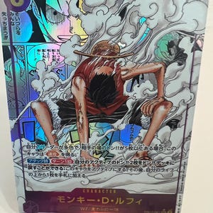May include: A holographic trading card featuring Monkey D. Luffy from the One Piece anime. The card shows Luffy in a dynamic pose, surrounded by smoke, with the number 6 and the text "7000" visible. The card has Japanese text.