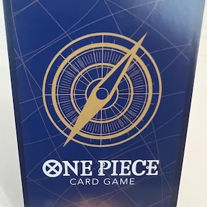 Puede incluir: Una tarjeta coleccionable azul con un dise&ntilde;o de br&uacute;jula dorada y una aguja larga. La tarjeta tiene las palabras "ONE PIECE CARD GAME" en texto blanco. La tarjeta est&aacute; sujeta por un soporte de pl&aacute;stico transparente.