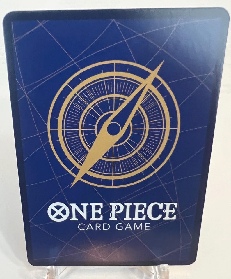 Puede incluir: Una tarjeta coleccionable azul con un dise&ntilde;o de br&uacute;jula dorada y una aguja larga. La tarjeta tiene las palabras "ONE PIECE CARD GAME" en texto blanco. La tarjeta est&aacute; sujeta por un soporte de pl&aacute;stico transparente.