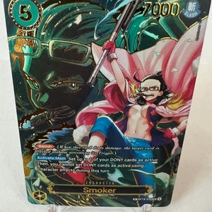 Puede incluir: Una tarjeta coleccionable con el personaje Smoker de la Marina Punk Hazard. La tarjeta tiene una combinación de colores verde y dorado con el número 5 en la esquina superior izquierda y el número 7000 en la superior derecha. El personaje lleva gafas, una capa rosa y pantalones azules.