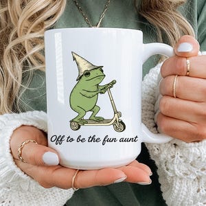 May include: White ceramic mug featuring a green frog riding a scooter and wearing a hat. The mug has the text "Off to be the fun aunt" in black script. The mug has a large handle.