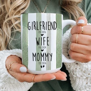 May include: White ceramic mug with green vertical stripes and the words "Girlfriend, Wife, Mommy" in black script. The mug includes the years 2016, 2024, and 2026, along with baby footprints. The mug is held by a person.