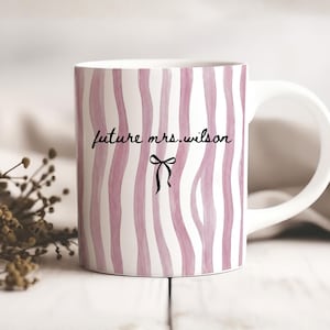 May include: A white ceramic mug with a black handle and the text "But First, Coffee" in a bold, black font. The mug is set against a blurred background, suggesting a cozy setting.