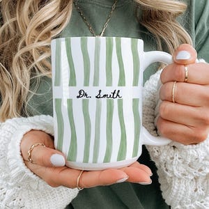 May include: White ceramic mug with a green and white striped pattern. The mug features the text "Dr. Smith" in black script. The mug has a white handle and is held by a person wearing a white sweater.