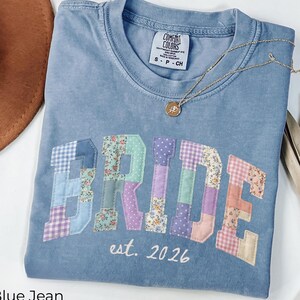 May include: A blue jean Comfort Colors t-shirt with the word "BRIDE" in patchwork letters. The patchwork includes floral and plaid patterns. The text "est. 2026" is embroidered below the word "BRIDE". A gold necklace with a pendant is on the shirt.