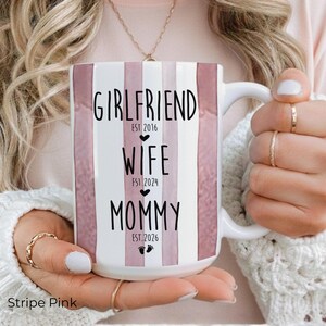May include: White ceramic mug with pink vertical stripes and the words "GIRLFRIEND EST 2016", "WIFE EST 2024", and "MOMMY EST 2026" in black. The mug has a white handle and is held by a person.