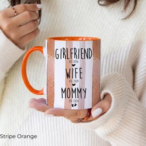 May include: An orange and white striped ceramic mug with an orange handle and rim. The mug features the words "Girlfriend," "Wife," and "Mommy" with the years 2016, 2024, and 2026 printed below each word. The mug is held by a person.