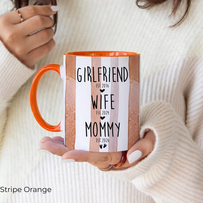 May include: An orange and white striped ceramic mug with an orange handle and rim. The mug features the words "Girlfriend," "Wife," and "Mommy" with the years 2016, 2024, and 2026 printed below each word. The mug is held by a person.