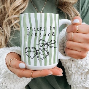 May include: White ceramic mug with green vertical stripes and the words "cheers to forever". The mug features a bow and two heart-shaped designs with the names "Kathryn" and "Grant".