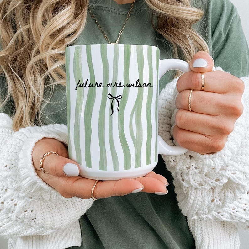 May include: White ceramic mug with a green and white striped pattern. The mug features the text "future mrs. wilson" in black script, with a black bow graphic below. The mug has a large handle and is held by a person.