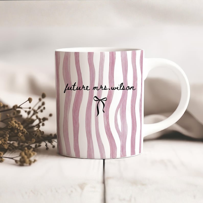 May include: A white ceramic mug with a black handle and the text "But First, Coffee" in a bold, black font. The mug is set against a blurred background, suggesting a cozy setting.