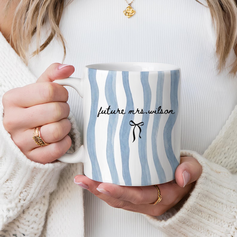 May include: White ceramic mug with blue wavy stripes and the text "future mrs. wilson" in black script, along with a black bow graphic. The mug has a white handle and is held in a person's hands.