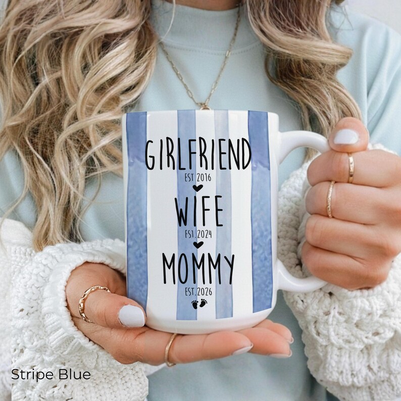 May include: White ceramic mug with blue vertical stripes. The mug features the words "Girlfriend EST 2016", "Wife EST 2024", and "Mommy EST 2026" in black text. The mug has a white handle and is held by a person.