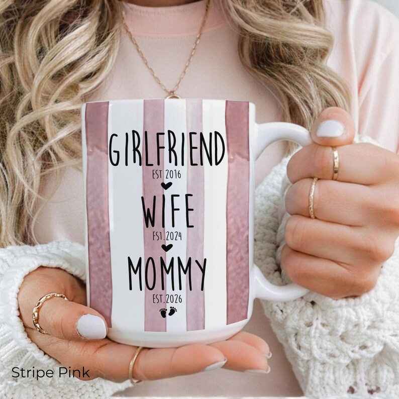 May include: White ceramic mug with pink vertical stripes and the words "GIRLFRIEND EST 2016", "WIFE EST 2024", and "MOMMY EST 2026" in black. The mug has a white handle and is held by a person.