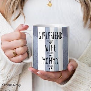 May include: White ceramic mug with blue and white vertical stripes. The mug features the words "Girlfriend," "Wife," and "Mommy" with corresponding dates. The mug is held by a person wearing a white sweater and gold jewelry.