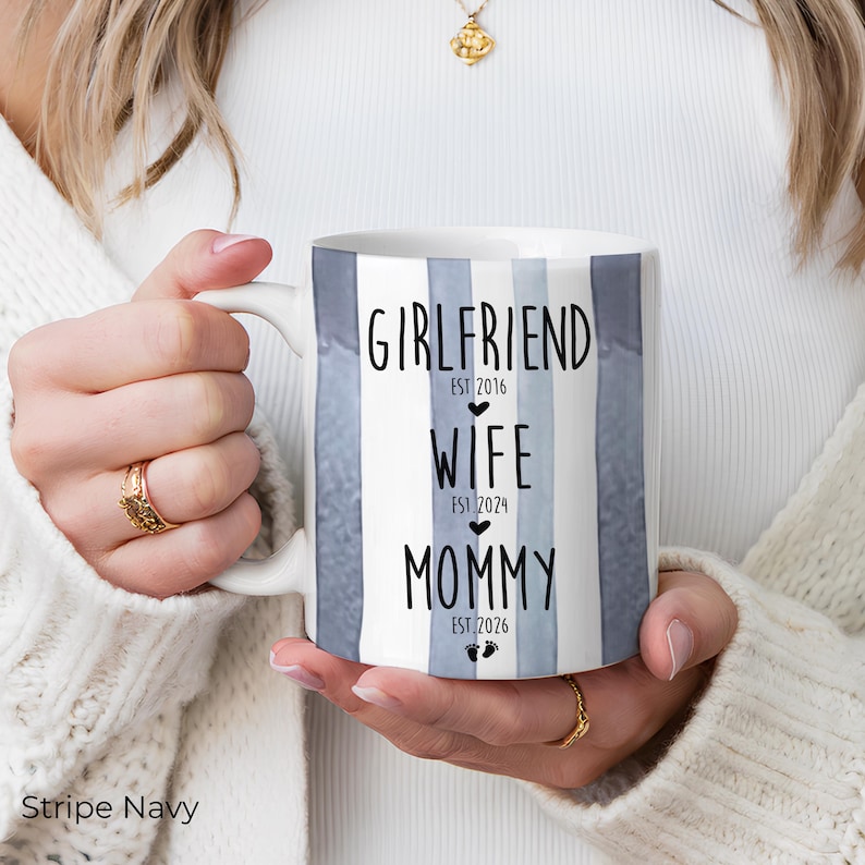 May include: White ceramic mug with blue and white vertical stripes. The mug features the words "Girlfriend," "Wife," and "Mommy" with corresponding dates. The mug is held by a person wearing a white sweater and gold jewelry.