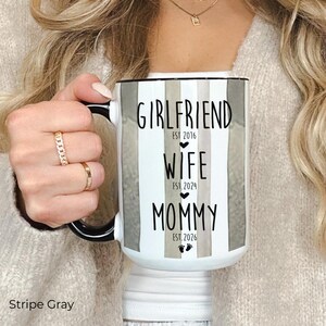 May include: White ceramic mug with black handle and rim, featuring gray and white vertical stripes. The mug has the words "GIRLFRIEND EST 2016", "WIFE EST 2024", and "MOMMY EST 2026" printed in black. Small footprints are at the bottom.