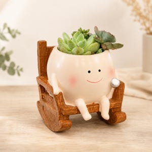 May include: A cream-colored ceramic planter shaped like a round character with a smiling face and pink cheeks, sitting in a brown textured rocking chair. The planter holds a green succulent plant and features a small silver-colored circular detail.