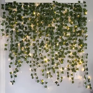 May include: A wall decoration featuring artificial green ivy vines with small, warm white LED lights. The vines cascade down, creating a natural, illuminated display. The lights are evenly spaced throughout the foliage.