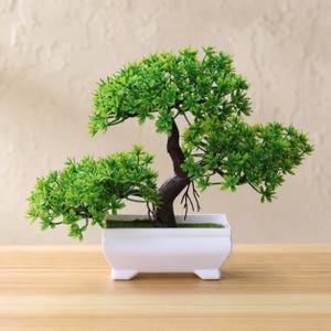Può includere: Un bonsai artificiale con fogliame verde brillante e un tronco marrone scuro testurizzato, piantato in un contenitore rettangolare in ceramica bianca con quattro piccoli piedini. La pianta presenta foglie dense e radici visibili.