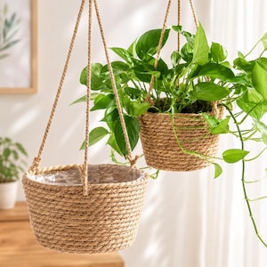 Pode incluir: Dois vasos suspensos feitos de corda natural enrolada em tom bege. Um vaso está vazio com um forro de plástico transparente visível no interior, enquanto o outro contém uma planta pothos verde exuberante com ramos pendentes.