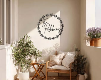 May include: A black metal family name sign, featuring the name "The Robbins" in elegant script within a circular wreath of leaves. The sign is mounted on a light-colored wall, enhancing its visibility. The scene includes a wooden chair, plants, and a small table.