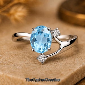 May include: A silver-toned ring featuring a large oval-cut light blue gemstone as the centrepiece, accented by two small round-cut clear stones on the split band. The text TheCypherCreation is visible at the bottom of the image.