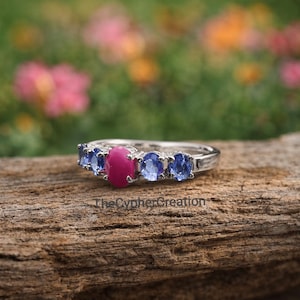Puede incluir: Un anillo de plata con una piedra central de rubí ovalada flanqueada por cuatro zafiros azules ovalados. El anillo está sobre una superficie de madera con un fondo floral borroso. El texto "TheCypherCreation" es visible.