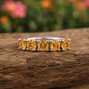 Può includere: Un anello color argento con cinque pietre preziose rotonde di colore arancione brillante disposte in fila. La fascia è metallica e lucida, e il testo TheCypherCreation è visibile sulla superficie sotto l'anello. Le pietre sono uniformi.