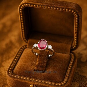 Puede incluir: Anillo de plata con una gran piedra central ovalada de rubí y dos piedras blancas más pequeñas a cada lado. El anillo se muestra en un joyero de terciopelo marrón. El rubí es de color rojo intenso.