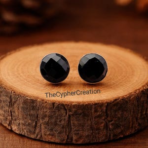 Puede incluir: Un par de pendientes de botón negros y facetados con un engaste plateado. Los pendientes son redondos y están sobre una superficie de madera. El texto "TheCypherCreation" es visible en la madera.