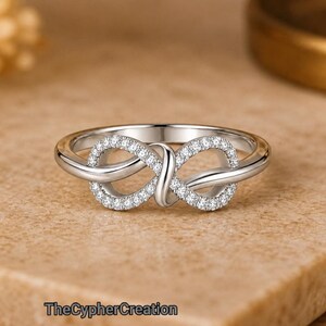 Puede incluir: Un anillo de tono plateado con un símbolo de infinito como pieza central. El bucle del infinito está incrustado con pequeñas piedras claras y brillantes a lo largo de su superficie. La banda es lisa y pulida, conectándose al diseño central.
