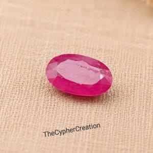 Pode incluir: Uma gema rosa vibrante, de formato oval e facetada com uma superfície polida. A pedra apresenta um tom magenta profundo e facetas geométricas claras que refletem a luz. O texto TheCypherCreation está impresso em preto abaixo da gema.