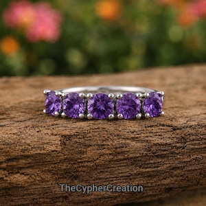 Op de afbeelding: Een zilverkleurige ring met vijf ronde paarse stenen op een rij. De band is van gepolijst metaal en houdt elke steen vast met kleine pootjes. De tekst TheCypherCreation is onderaan de afbeelding zichtbaar in een donker lettertype.