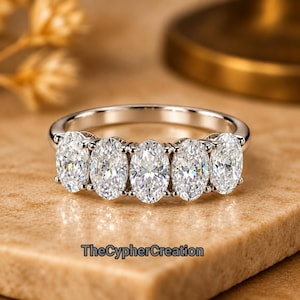 Peut inclure: Une bague argentée ornée de cinq pierres claires taille ovale disposées en rangée sur le dessus. L'anneau est lisse et poli. Le texte TheCypherCreation est visible en lettres noires au bas de l'image, sous la bague.