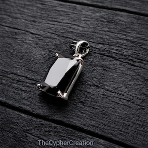 Op de afbeelding: Een rechthoekige zwarte hanger met een zilverkleurige zetting. De hanger is geplaatst tegen een donkere achtergrond van houtnerf. TheCypherCreation staat onderaan de afbeelding.