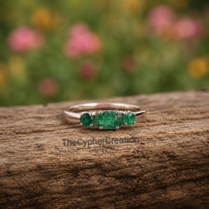 Peut inclure: Une bague en argent ornée de trois émeraudes vertes éclatantes. L'émeraude centrale est rectangulaire, flanquée de deux émeraudes rondes. La bague est posée sur une surface en bois rustique avec un arrière-plan floral flou.