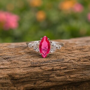 Puede incluir: Un anillo de plata con una llamativa gema rosa brillante en forma de marquesa en el centro. La piedra principal está flanqueada por piedras más pequeñas y claras en forma de marquesa. El anillo está sobre un fondo natural borroso.