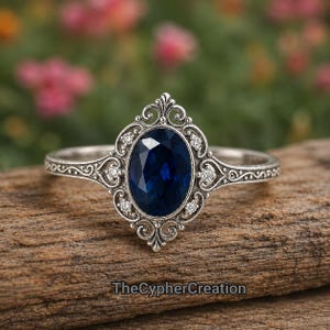 Puede incluir: Un anillo de plata con una piedra preciosa azul oscuro de corte ovalado en el centro, rodeada por un marco de filigrana decorativo con pequeñas piedras claras. La banda muestra intrincados patrones de metal y el texto TheCypherCreation.