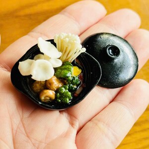 May include: Miniature black bowl with a lid, filled with a detailed food arrangement. The food includes white mushrooms, green vegetables, and other colorful ingredients. The bowl is held in a hand against a wooden background.