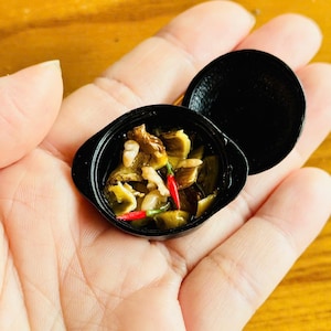 May include: Miniature black pot filled with a dish of food, including what appears to be mushrooms, green vegetables, and red peppers. The pot is open, revealing the contents. The pot is held in a hand against a wooden background.