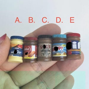 May include: A selection of miniature food jars, featuring labels such as "Kraft Mayo" and "Lotus." The jars have different coloured lids and labels. The jars are held in a hand.