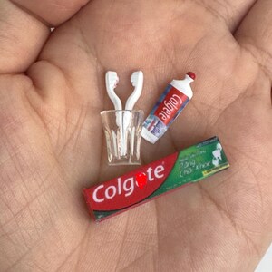 May include: Miniature dental hygiene set including a clear glass with two white toothbrushes, a tube of Colgate toothpaste, and a toothpaste box. The Colgate products have red and white branding.
